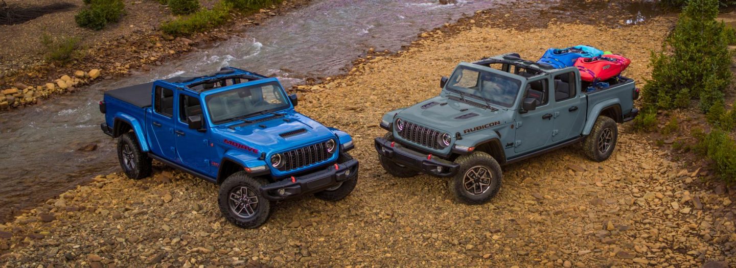 A blue Jeep Gladiator Mojave exiting a stream and a gray Gladiator Rubicon parked nearby, both with their tops removed.