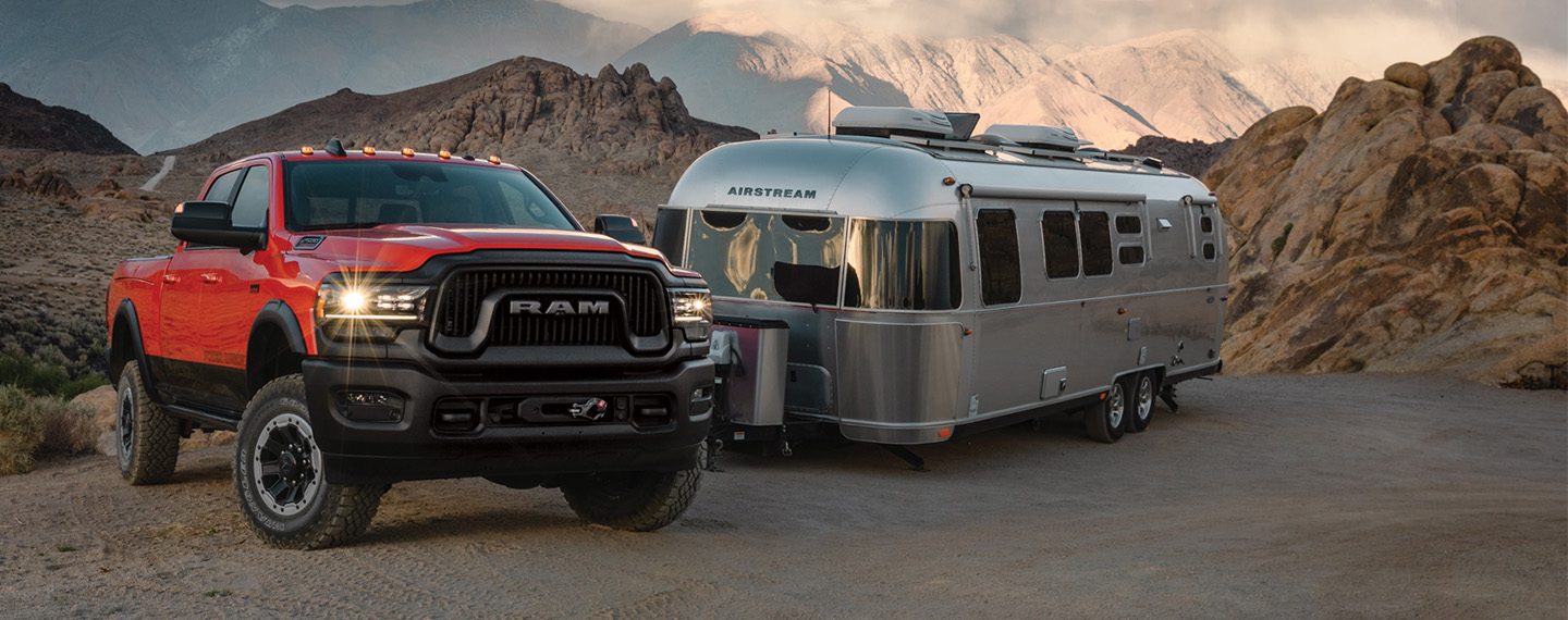 A red 2022 Ram 2500 Power Wagon with its headlamps on, parked off-road next to a travel trailer, on a clearing in the mountains.