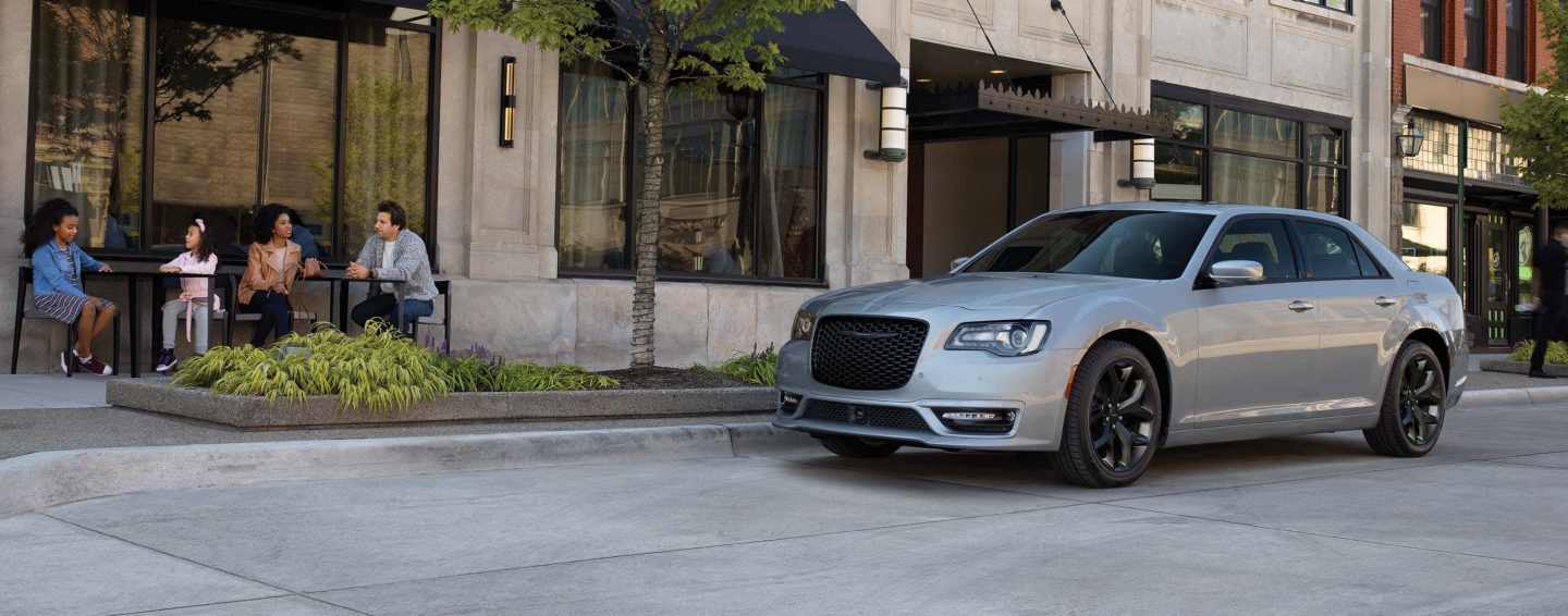 A silver 2023 Chrysler 300 TBD parked at the curb beside a sidewalk cafe.
