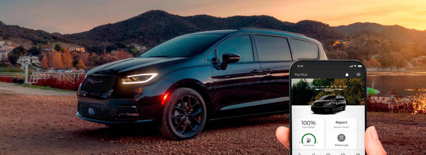 In the foreground, a smartphone displaying a page from the Chrysler app. In the background a black Chrysler Pacifica parked beside a lake.