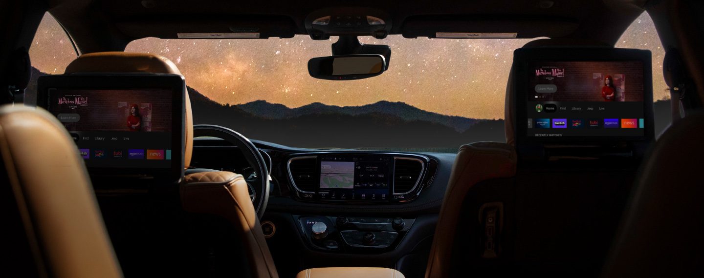 Interior image of an empty vehicle from the backseat looking forward. A navigation map is visible on the vehicle’s touchscreen. Through the windshield, a starry night sky can be seen above tree-covered ridgelines.